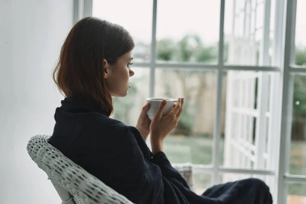 Женщина с чашкой чая сидит в кресле, покрытом одеялом, и смотрит в окно — стоковое фото