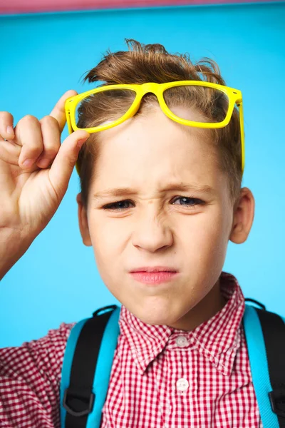 Écolier en lunettes jaunes avec un sac à dos et une chemise rose sur fond bleu — Photo