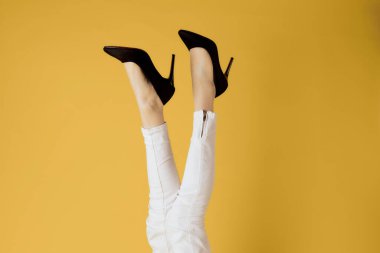 Kadınların ters bacakları ve siyah ayakkabıları. Beyaz kot, izole edilmiş sarı arka plan.