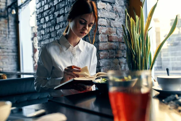 Mulher feliz na camisa e saia vermelha senta-se a uma mesa em um café com bloco de notas e caneta na mão — Fotografia de Stock