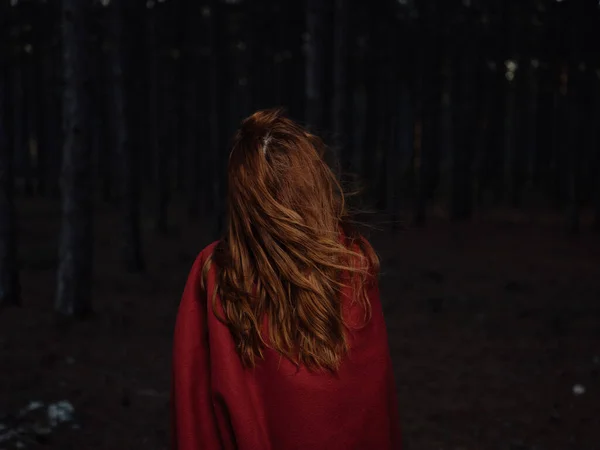 Una mujer viaja en el bosque por la noche con una tela escocesa roja sobre sus hombros, vista hacia atrás —  Fotos de Stock