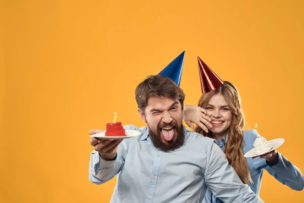 Aniversário festa homem e mulher em um fundo amarelo em chapéus com um bolo em suas mãos — Fotografia de Stock