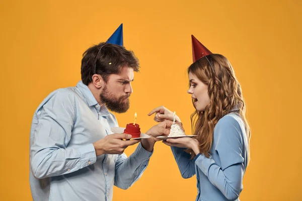 Sarı arka planda pastası olan şapkalı kadın ve erkek doğum günü partisi. — Stok fotoğraf