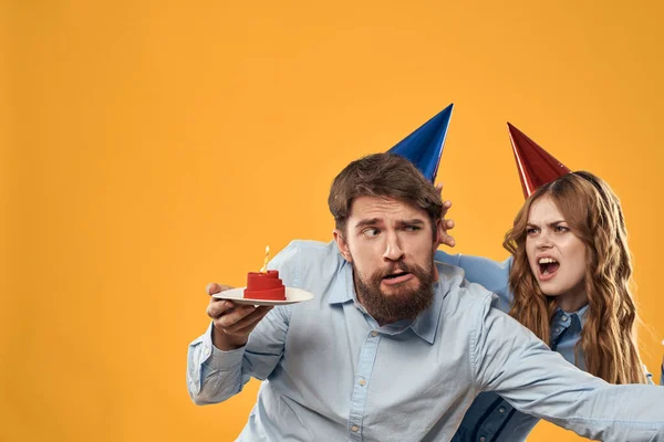 노란색 배경에 손에 케이크를 들고 모자를 쓴 생일 파티를 하는 남자와 여자 — 스톡 사진