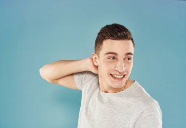 Sevimli adam elini başının arkasında tutuyor. Duyguları var. Mavi arka plan beyaz tişört.