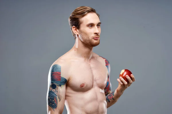 Atleta sexy con músculos bombeados come una manzana sobre un fondo gris y un tatuaje en su brazo — Foto de Stock