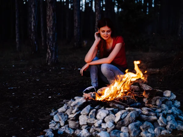 Una mujer se sienta cerca del fuego y mira el fuego en un bosque de pinos —  Fotos de Stock