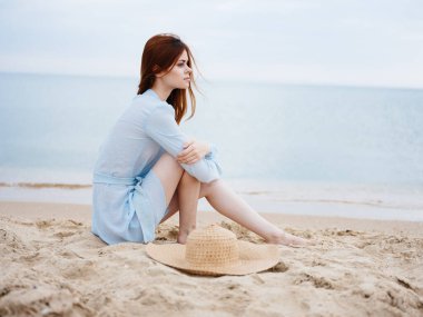 Romantik bir kadın oturmuş şapkası ve mavi elbisesi kumlu denizle