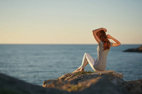 Kobieta w spodniach i koszulce siedzi na wielkim kamieniu nad morzem z rękami w górze. — Zdjęcie stockowe