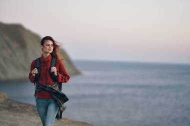 Sırt çantalı bir kadın yürüyüşçü dağlarda deniz kenarında yürüyor.