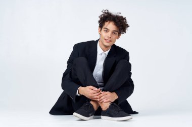 Takım elbiseli şık bir genç adam parlak bir odada bacak bacak üstüne atmış şekilde yerde oturuyor.