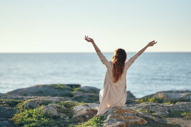Kadın kazak giyer, ellerini başının üstüne koyar. Deniz kıyısındaki dağlarda. Temiz hava.