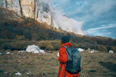 Kafasında sırt çantası olan, mavi gökyüzü ve yüksek dağları olan ceketli bir kadının yolculuğu.