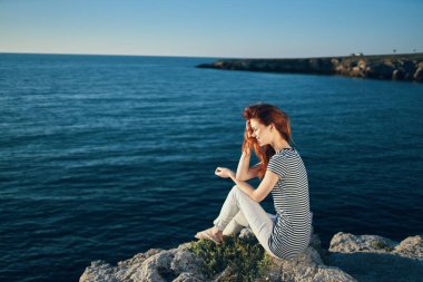 Tişörtlü ve pantolonlu bir kadın deniz manzaralı sahilde bir kayanın üzerinde oturuyor.