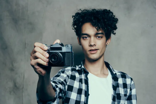 Ung man med en kamera i handen och i en rutig t-shirt på en grå bakgrund inomhus fotograf — Stockfoto