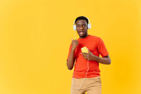 Afro-americano com um telefone em suas mãos em fones de ouvido escuta música fundo amarelo — Fotografia de Stock