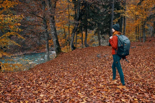 Vrouw in de herfst in het park met gevallen bladeren en een rugzak op haar rug rivier op de achtergrond — Stockfoto