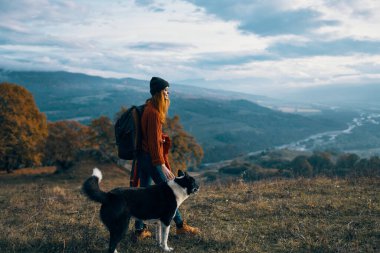 Sırt çantalı bir kadın, köpek yürüyüşünün yanında, dağ manzarası.