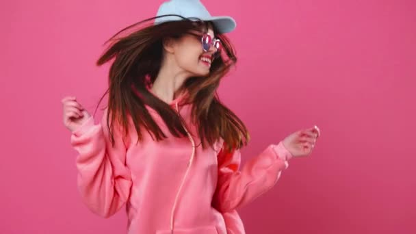 Mulher de boné e óculos dançando alegremente em fundo rosa — Vídeo de Stock