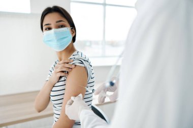 Beyaz önlüklü bir doktor, hastane kovid aşısı olan bir kadının omzuna iğne yapar.