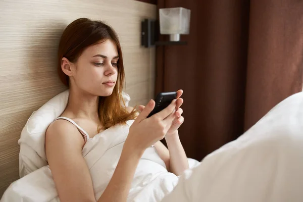 Mulher com telefone móvel encontra-se na cama perto da janela e cortinas design — Fotografia de Stock