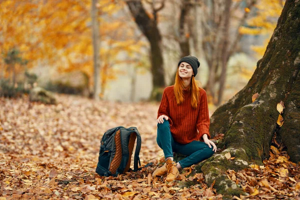 Kobieta w spodniach i butach z plecakiem siedzi w pobliżu drzewa w jesiennym lesie spadły liście — Zdjęcie stockowe
