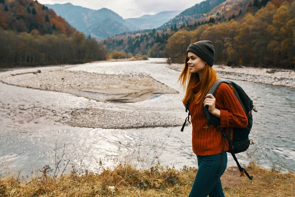 Una mujer con una chaqueta roja y una mochila en la espalda están caminando a orillas del río en las montañas —  Fotos de Stock