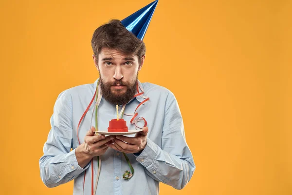 Hombre alegre con un pastel en un fondo amarillo cumpleaños días festivos gorra en su cabeza —  Fotos de Stock
