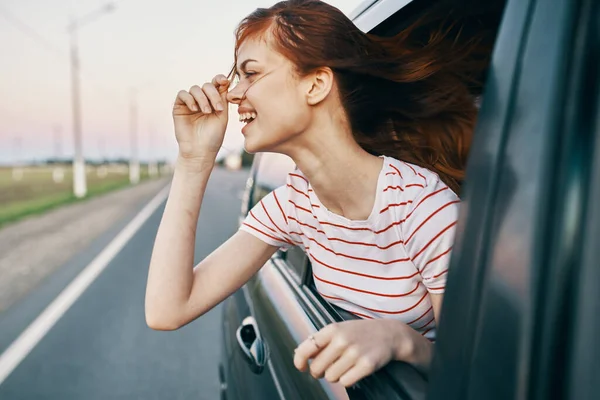 Mujer pelirroja en una camiseta mira por la ventana del coche y hace gestos con las manos en la carretera de verano — Foto de Stock