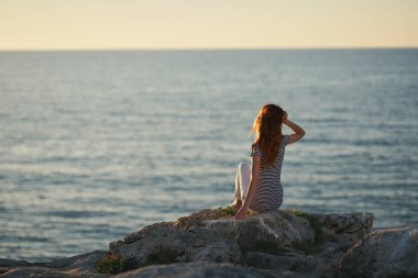Bir kadın denizin yanındaki büyük taşın üzerinde oturur ve dağlarda gün batımını seyreder.
