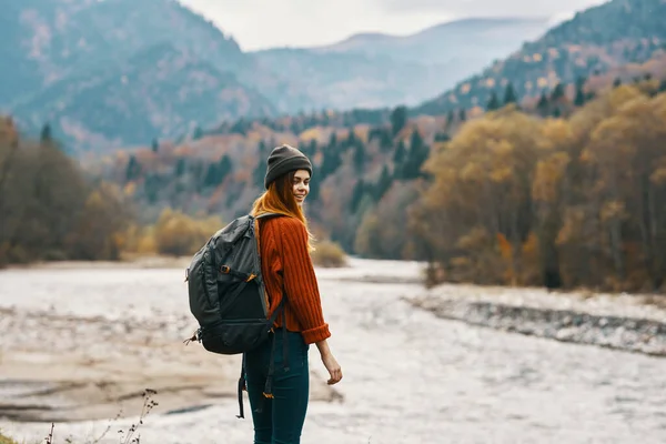 Mulher em um boné camisola vermelha com uma mochila na margem do rio e montanhas na distância — Fotografia de Stock