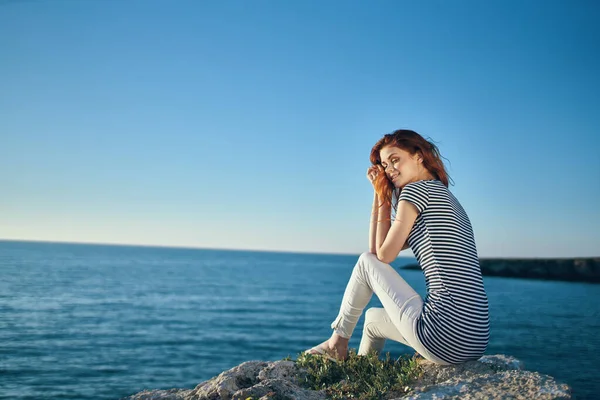 Donna in t-shirt a righe e pantaloni bianchi in estate su una pietra vicino al mare in montagna — Foto Stock