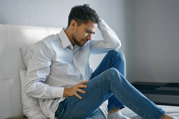 A lonely man in a shirt and jeans sits on the bed and holds his head with his hand