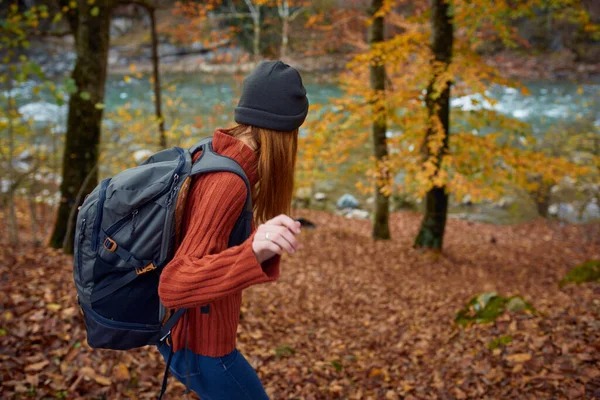 Žena ve svetru s batohem na zádech v blízkosti řeky v horách a parku stromy podzimní krajiny — Stock fotografie