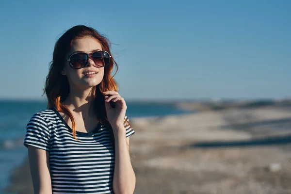 Kobieta w okularach przeciwsłonecznych i w pasiastym t-shircie gesty dłońmi w górach nad morzem na plaży — Zdjęcie stockowe