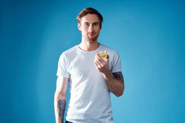 Χαριτωμένος τύπος με τατουάζ και λευκό μπλουζάκι σε μπλε φόντο που τρώει μήλα. — Φωτογραφία Αρχείου