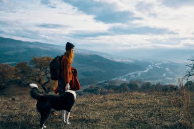 Sırt çantalı bir kadın, köpek yürüyüşünün yanında, dağ manzarası.
