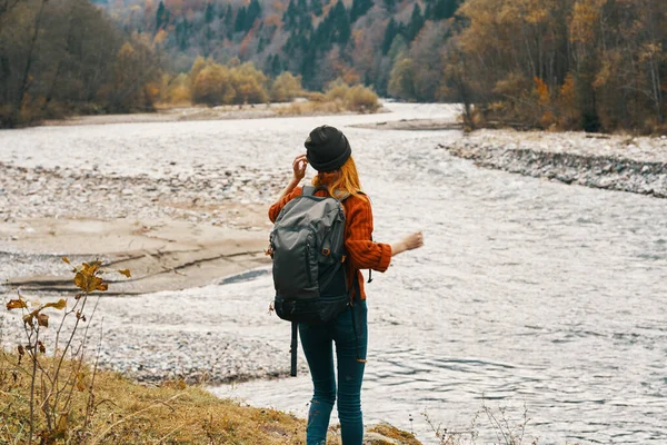 Une voyageuse en pull avec un sac à dos fait des gestes avec ses mains et se détend sur la rive de la rivière dans les montagnes — Photo