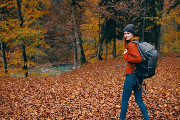 Frau mit Hut, rotem Pullover und Jeans spaziert mit Rucksack auf dem Rücken durch den Park — Stockfoto