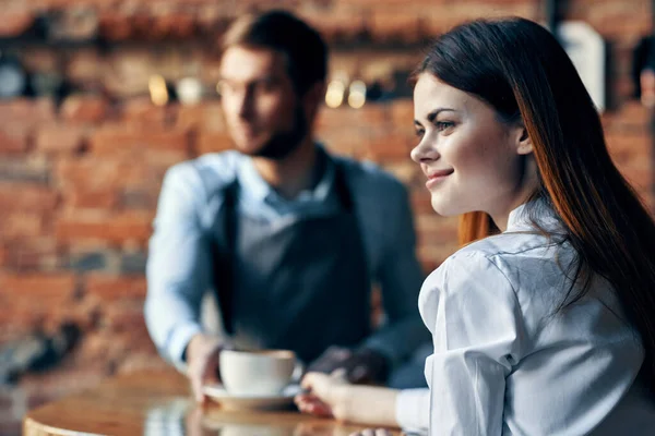 Garçom em avental cinza senta-se à mesa e cliente com uma xícara de parede de tijolo de café modelo interior — Fotografia de Stock