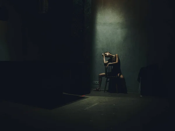 Une femme s'assoit sur une chaise avec les jambes écartées dans une pièce sombre et une lumière tombante d'en haut — Photo
