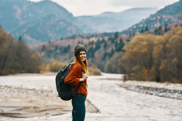 woman in a red sweater cap with a backpack on the river bank and mountains in the distance
