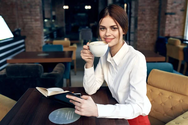 Bela mulher com uma xícara na mão senta-se a uma mesa em um café e um livro em sua mão modelo de restaurante — Fotografia de Stock