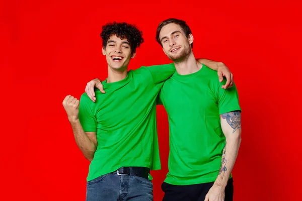 Lustige Freunde grüne T-Shirts Umarmungen Emotionen Freude roter Hintergrund — Stockfoto