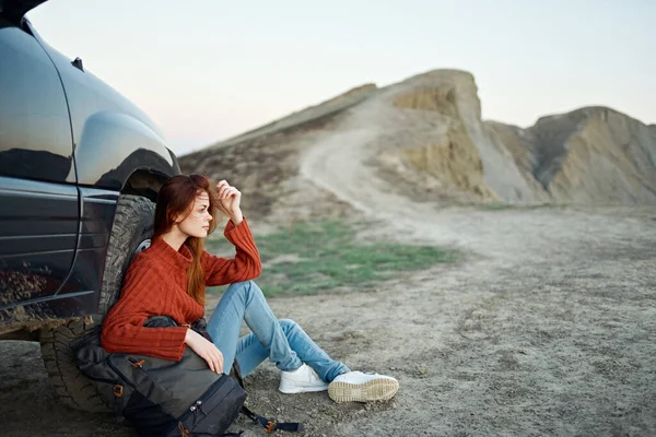 Um viajante em uma camisola e jeans senta-se no chão com uma mochila na mão perto de um carro nas montanhas — Fotografia de Stock