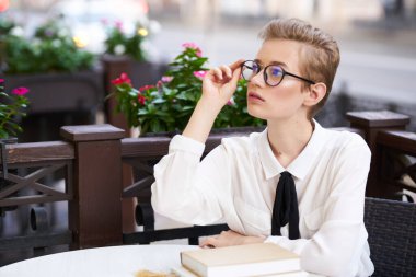 Kafedeki bir masada kitap taşıyan gözlüklü ve gömlekli genç bir kadın.