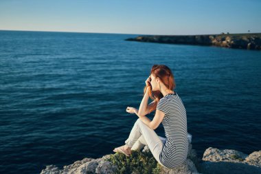 Tişörtlü ve pantolonlu bir kadın deniz manzaralı sahilde bir kayanın üzerinde oturuyor.