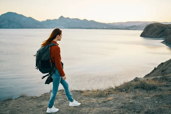 Sırt çantalı, kot pantolonlu ve spor ayakkabılı bir gezgin dağlarda deniz kenarında bir sahilde.