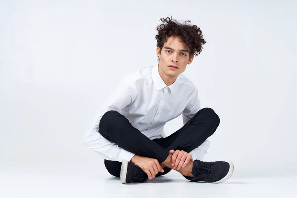 Um cara com cabelo encaracolado em uma camisa branca, tênis e calças senta-se no chão em uma sala brilhante — Fotografia de Stock