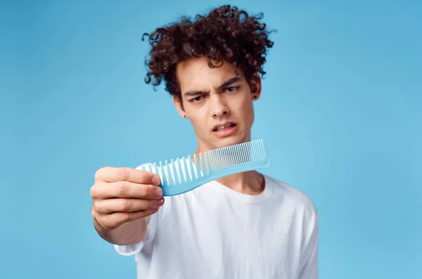 Cara com cabelo encaracolado segurando um pente na frente de seu rosto em um fundo azul e nojo — Fotografia de Stock